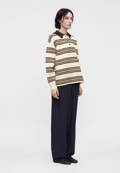Personne aux cheveux courts et foncés portant une chemise à manches longues beige et brune rayée, un pantalon noir large et des chaussures noires, se tenant de côté.