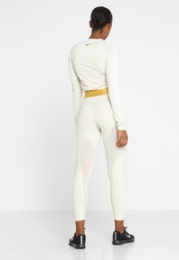 Top de manga larga beige claro con un patrón texturizado, combinado con leggings blancos lisos que tienen paneles de malla y una cinturilla dorada. Zapatillas negras.