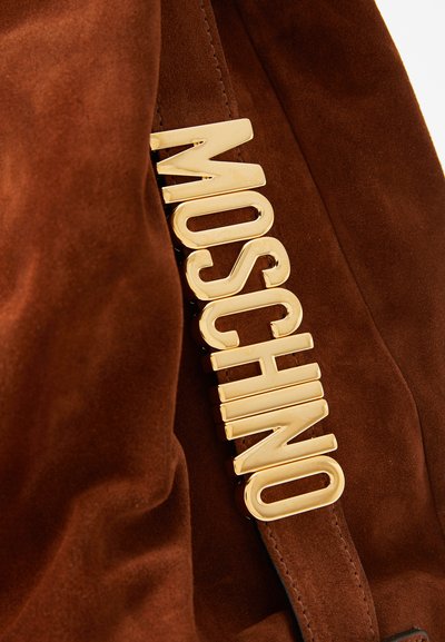 Brun mockaskinnväska med en framträdande guldfärgad "Moschino"-logotyp med en slät yta. Texturen är mjuk och fyllig.