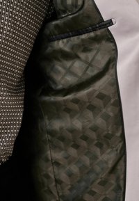 Blazer mit einem gemusterten Stoff mit geometrischen Formen in Schwarz und Weiß, der kontrastiert mit einem glatten, dunkelgrünen Futter und schwarzen Akzenten.