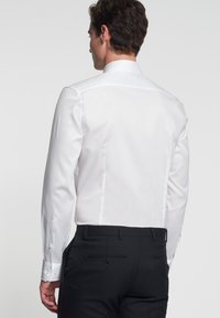 Chemise blanche à manches longues et poignets boutonnés, associée à un pantalon noir. Texture lisse, design ajusté, lignes épurées et détails minimalistes.
