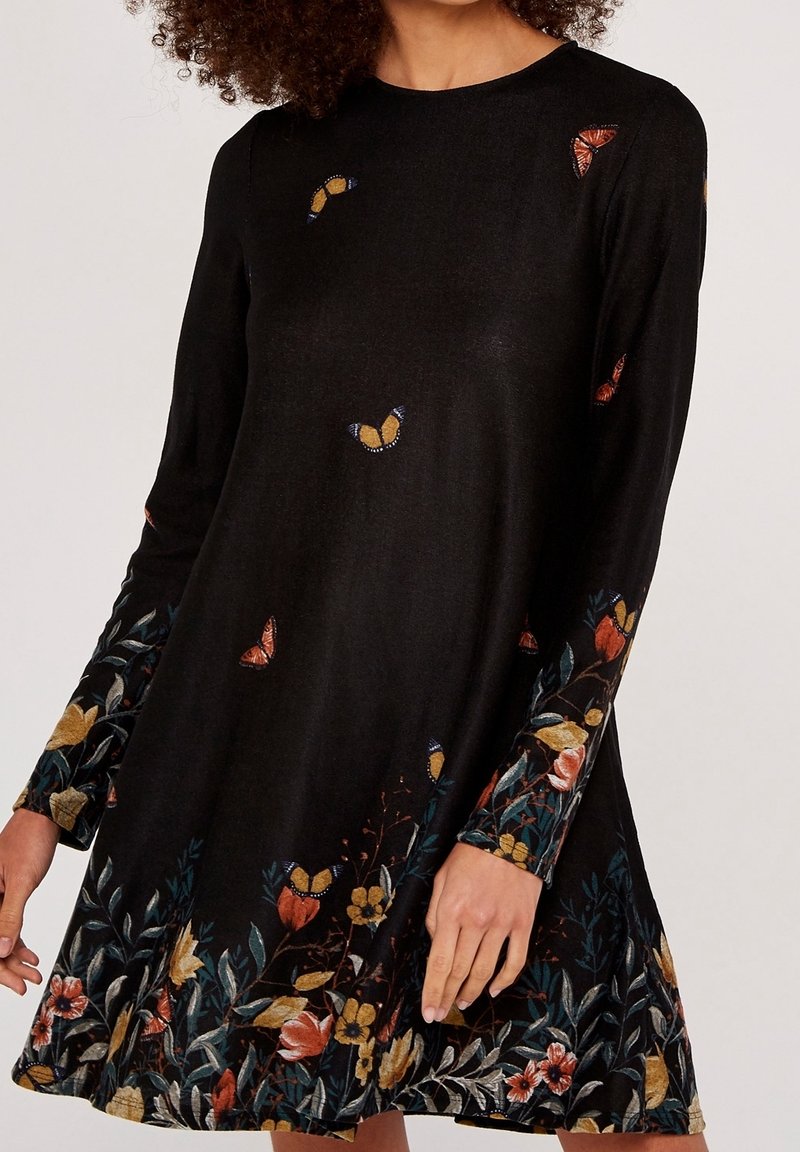 Robe noire à manches longues avec des motifs colorés de papillons et de fleurs le long des manches et de l'ourlet portée par une femme.