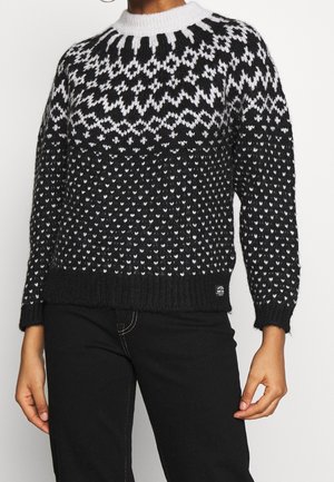 Pull en maille à motif noir et blanc avec un col montant côtelé, associé à un pantalon noir avec des coutures blanches visibles.