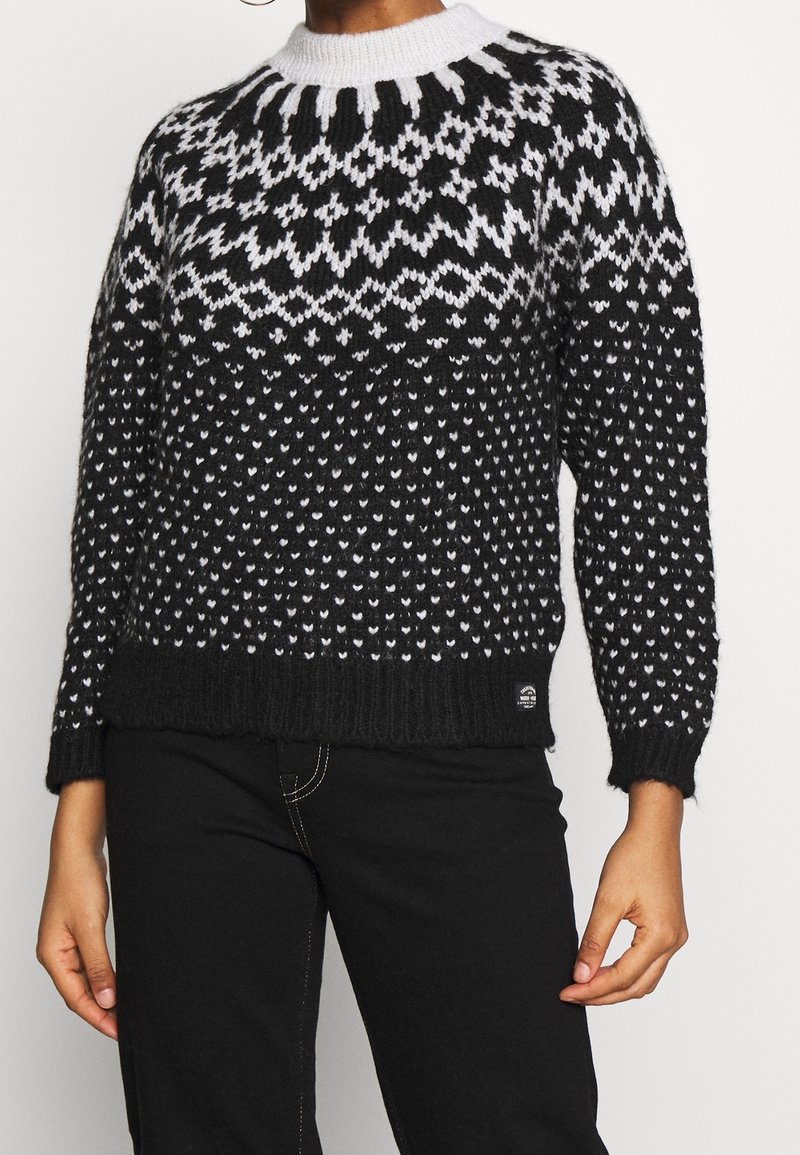 Pull en maille à motif noir et blanc avec un col montant côtelé, associé à un pantalon noir avec des coutures blanches visibles.