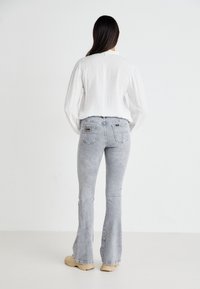 Jeans grigi chiari a gamba svasata con una texture sbiadita, dotati di tasche posteriori e un'etichetta con logo, abbinati a una blusa bianca con texture.