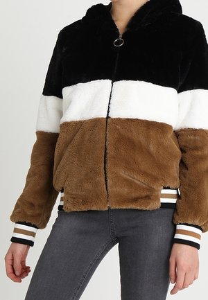 Femme portant une veste duveteuse à blocs de couleur noir, blanc et marron avec des poignets rayés et un jean gris foncé.