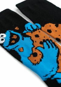 Calzini neri con un personaggio dei cartoni animati blu che tiene un biscotto, evidenziati da accenti arancioni e marroni. Design lavorato a maglia con motivi giocosi.