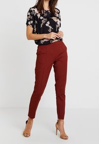 Haut noir à motifs floraux à manches courtes associé à un pantalon slim bordeaux. Le mannequin porte des sandales à talons hauts couleur tan, soulignant l'élégance de la tenue.