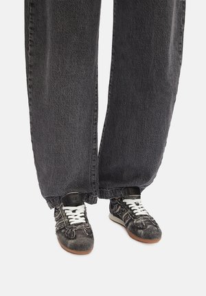 Stopy w czarnych trampkach z denimu z białymi sznurówkami oraz szerokich szarych spodniach o szerokich nogawkach na białym tle.