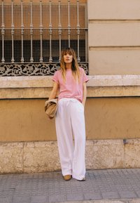 Femme debout contre un mur en pierre beige sous une grille de fenêtre en fer décorative, portant une chemise à rayures rouges, un pantalon blanc et tenant un chapeau beige.