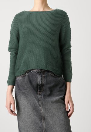 Grüner, strukturiert gestrickter Pullover mit weitem Ausschnitt und langen Ärmeln, kombiniert mit einem grauen Jeansrock mit geradem Schnitt und sichtbaren Nähten.