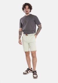 T-shirt grigia, pantaloni corti verde chiaro, scarpe da ginnastica marroni. I pantaloni corti presentano un orlo arrotolato e una cintura intrecciata; tatuaggi visibili sul braccio. Design semplice.