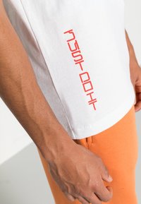 Vit bomull t-shirt med en röd vertikal "Just Do It"-tryck på vänster ärm, tillsammans med orangea byxor. Slät textur och avslappnad passform.