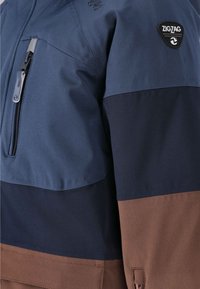 En flerfärgad jacka med navy, mörkblå och bruna sektioner, med en ficka med dragkedja och ett triangulärt logotypmärke på axeln.