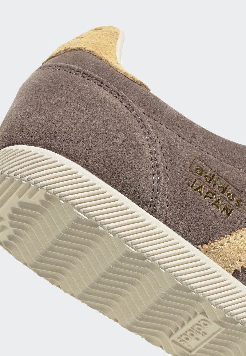 Grå mockasinsneaker med beige detaljer, texturerad vit gummisula och guldfärgad "adidas JAPAN"-logotyp. Har sömdekor på ovandelen.