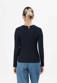 Frau mit blonden Haaren in einem tiefen Dutt, die ein enganliegendes schwarzes Rippenoberteil mit langen Ärmeln und blaue Jeans trägt, von hinten gesehen.