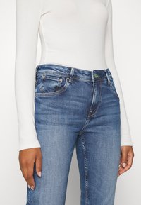 Blå denimjeans med en klassisk passform, med medelhög midja, femfickorsdesign och grön accentknapp. Slät textur med lätt urtvättning.