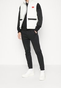 Weißer gepolsterter Weste mit schwarzen Akzenten, zwei Reißverschlusstaschen, kombiniert mit schwarzen Sweatpants und weißen Sneakers. Minimalistisches Design und lässige Passform.