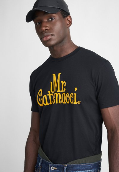 T-shirt en coton noir avec le texte en jaune vif "Mr. Catenacci" sur la poitrine. Associé à un pantalon en denim et une ceinture vert olive.