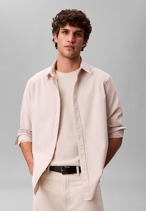 Junger Mann mit lockigem Haar, der ein hellbeiges Hemd mit Knöpfen über einem strukturierten cremefarbenen Pullover, helle Hose und schwarzen Gürtel trägt, Hände in den Taschen.