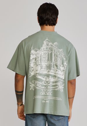 Uomo che indossa una T-shirt verde salvia con grafica bianca di un'auto d'epoca e un edificio storico, abbinata a jeans blu, mostrato di spalle.