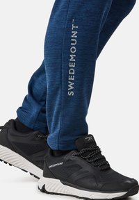 Blaue Heather-Jogginghosen mit silbernem "SWEDEMOUNT"-Logo an der Seite, kombiniert mit schwarzen Turnschuhen mit weißer Sohle und strukturiertem Design.