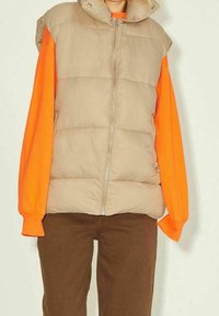 Beige vadderad väst med hög krage och dragkedja, buren över en livfull orange långärmad skjorta. Bruna byxor fulländar outfiten.