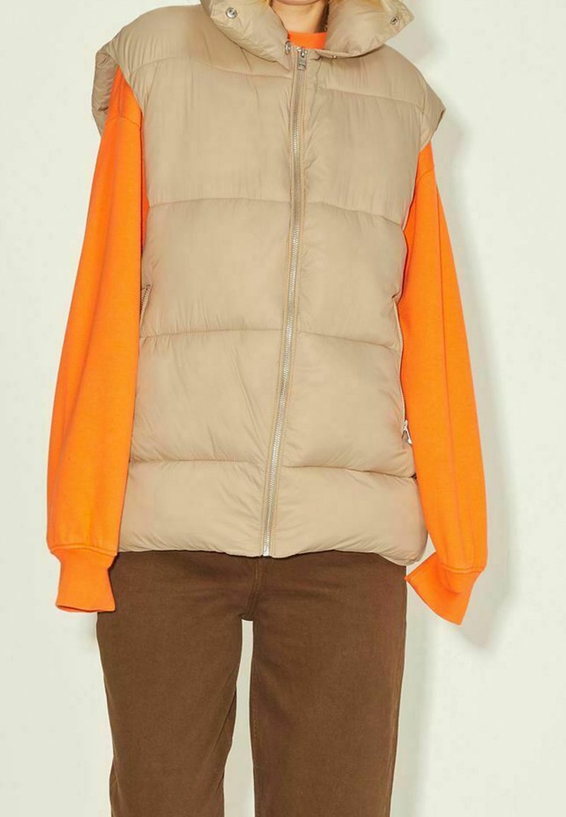 Beige vadderad väst med hög krage och dragkedja, buren över en livfull orange långärmad skjorta. Bruna byxor fulländar outfiten.