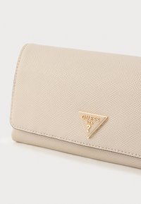 Beige színű, texturált pénztárca háromszög alakú arany Guess logóval. Hajtható kialakítású, az éleken elegáns varrással.