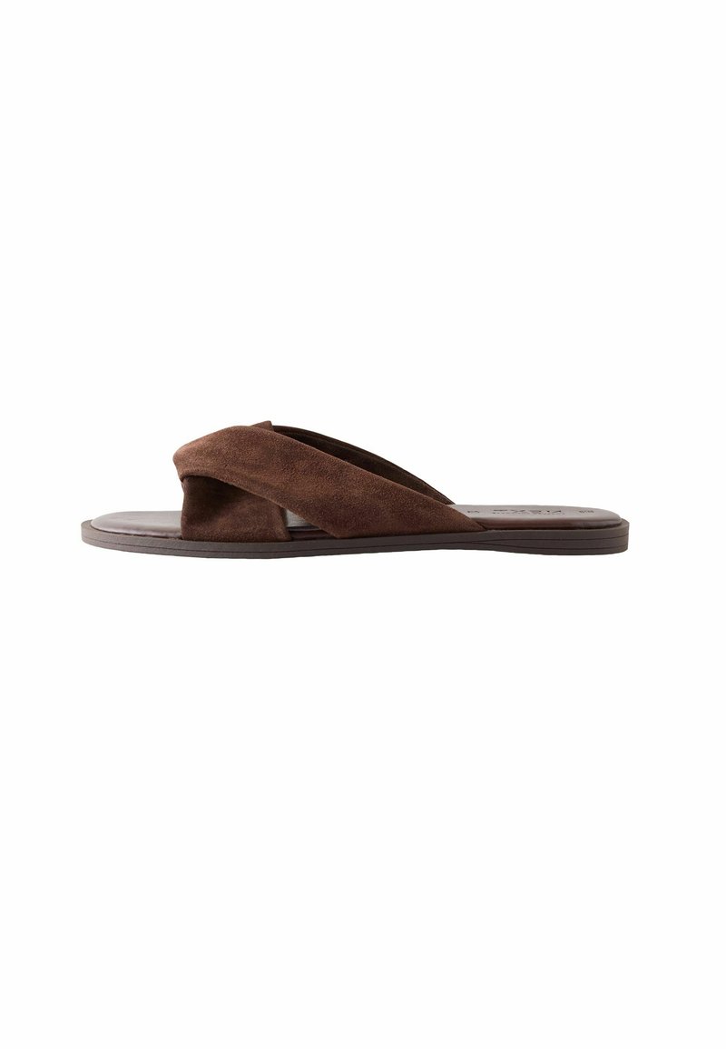 Bruine suède slide sandalen met kruisende brede banden en een platte zool, getoond in zijaanzicht op een witte achtergrond.