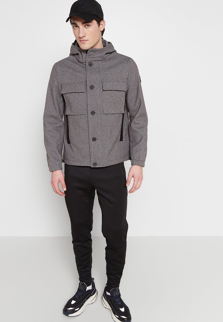 Grijze, gestructureerde jack met een capuchon, twee voorzakken en een knoopsluiting. Gepaard met zwarte, tapered broeken en zwarte sneakers.