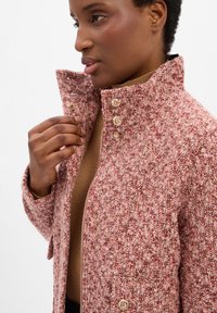 Strukturierte Jacke aus pinkem und rotem Tweed mit hohem Kragen, goldenen Knopfakzenten und einem Reißverschluss. Die Hand des Models befindet sich in der Nähe des Kragens.