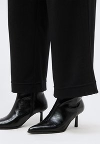 Μαύρες γυναικείες αστραγάλες με μυτερή μυτερή μύτη και λεπτά τακούνια, φορεμένες με μαύρο παντελόνι cropped, πάνω σε απλό φόντο.