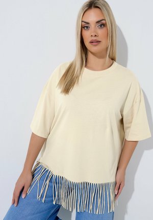 Femme blonde portant un T-shirt beige oversize avec un ourlet à franges, associé à un jean bleu, debout sur un fond gris clair uni.