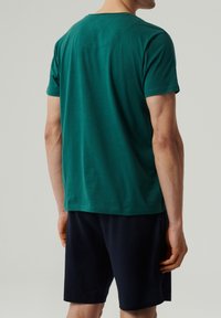 Zielona bawełniana koszulka z krótkim rękawem i okrągłym dekoltem, prezentowana na modelu w granatowych szortach. Gładka tekstura, luźny krój.