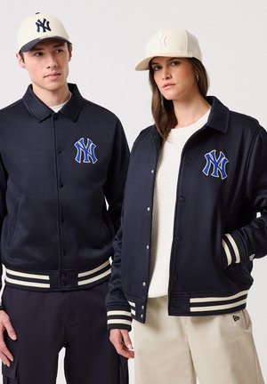 Deux mannequins portant des vestes bleu marine des New York Yankees et des casquettes claires, debout devant un fond uni.