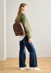Sac à dos en cuir marron avec une surface lisse, une forme arrondie, des accents dorés sur la fermeture éclair, et une veste verte matelassée portée par-dessus une chemise blanche.