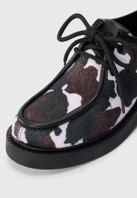 Chaussure en cuir noir et blanc avec un motif en cuir de vache, ornée d'accents verts et marron, bout rond et lacets noirs. Surface texturée.