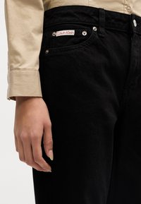 Mustat farkut, joissa on Calvin Kleinin logopatch, hopea varusteet ja lähikuva kädestä, joka lepää reisi-alueella.
