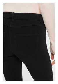 Gros plan d'une personne portant un pantalon noir ajusté et un haut à manches longues côtelé rose clair, montrant la zone de la poche arrière.