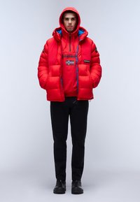 Giacca a vento rossa con cappuccio, presenta un rivestimento blu, chiusura con zip e toppe. Indossata sopra una felpa rossa, abbinata a pantaloni e scarpe nere.