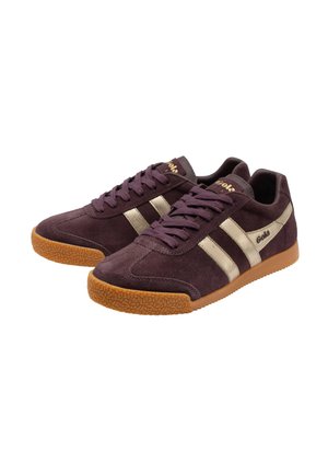 Een paar donkerpaarse suède sneakers met gouden strepen, gum rubberzolen en donkerpaarse veters, schuin naast elkaar geplaatst.
