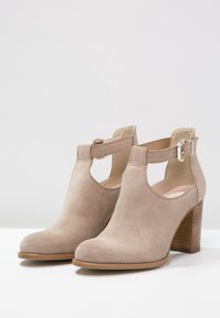 Beige mockaskinankelstövlar med öppna skärningar, justerbart vristband med metallspänne och en staplad träklack.