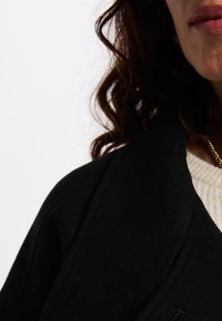 Cappotto in misto lana nero con superficie testurizzata, dotato di grandi rever e dettagli cuciti. Un maglione di lana crema è visibile sotto.