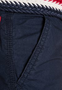 Námořnická modrá látka s texturovaným povrchem, včetně pleteného bílého a červeného pásku. Obsahuje boční kapsu s detailním šitím.