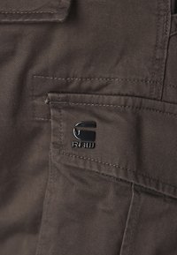 Bruine stoffen cargozak met een gestikte naad en een zwart metalen logodetail. Gladde textuur met een praktische uitstraling.