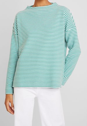 Persoon draagt een wit en turquoise horizontaal gestreept shirt met lange mouwen en witte broek, staand tegen een effen grijze achtergrond.