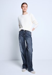 Suéter blanco esponjoso, jeans de tiro alto de color azul oscuro, calzado ligero. Modelo de pie con una mano en la cadera, fondo neutro.