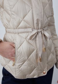 Giacca trapuntata color crema con cintura annodata in vita, tasca con cerniera dorata e bottone automatico, indossata sopra pantaloni scuri con una mano nella tasca.