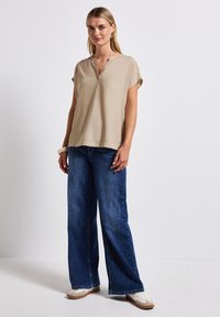 Beige Kurzarmoberteil mit V-Ausschnitt und lässiger Passform, kombiniert mit weit geschnittenen blauen Denim-Jeans und weißen Sneakers. Einfache, glatte Textur.